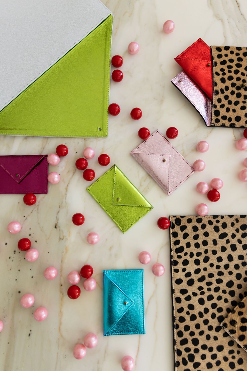 Collection of vibrant metallic envelope wallets in multiple colors on marble with red and pink decorative beads.