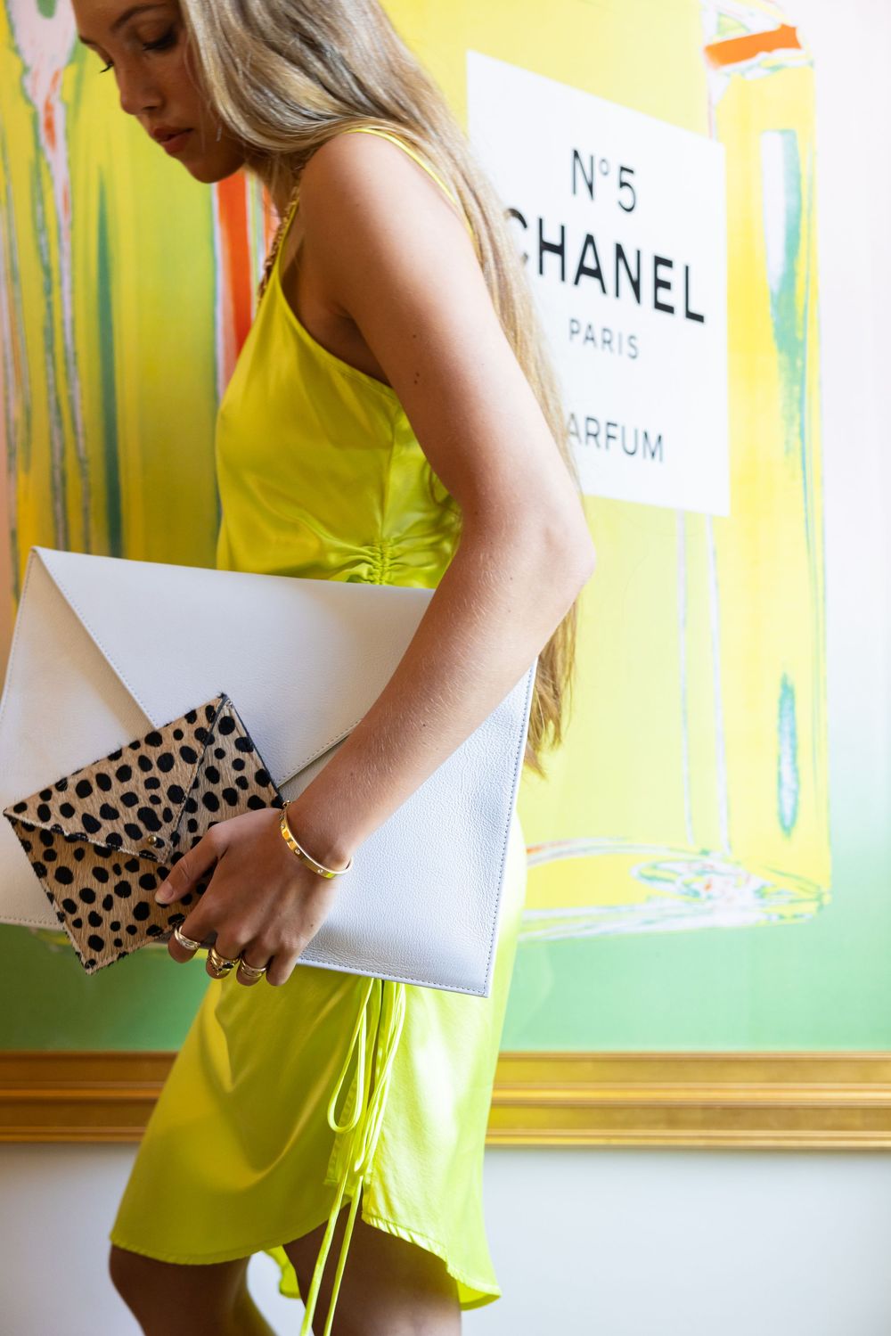 Blonde woman in neon yellow dress carries white envelope clutch and cheetah print bag near Chanel art.