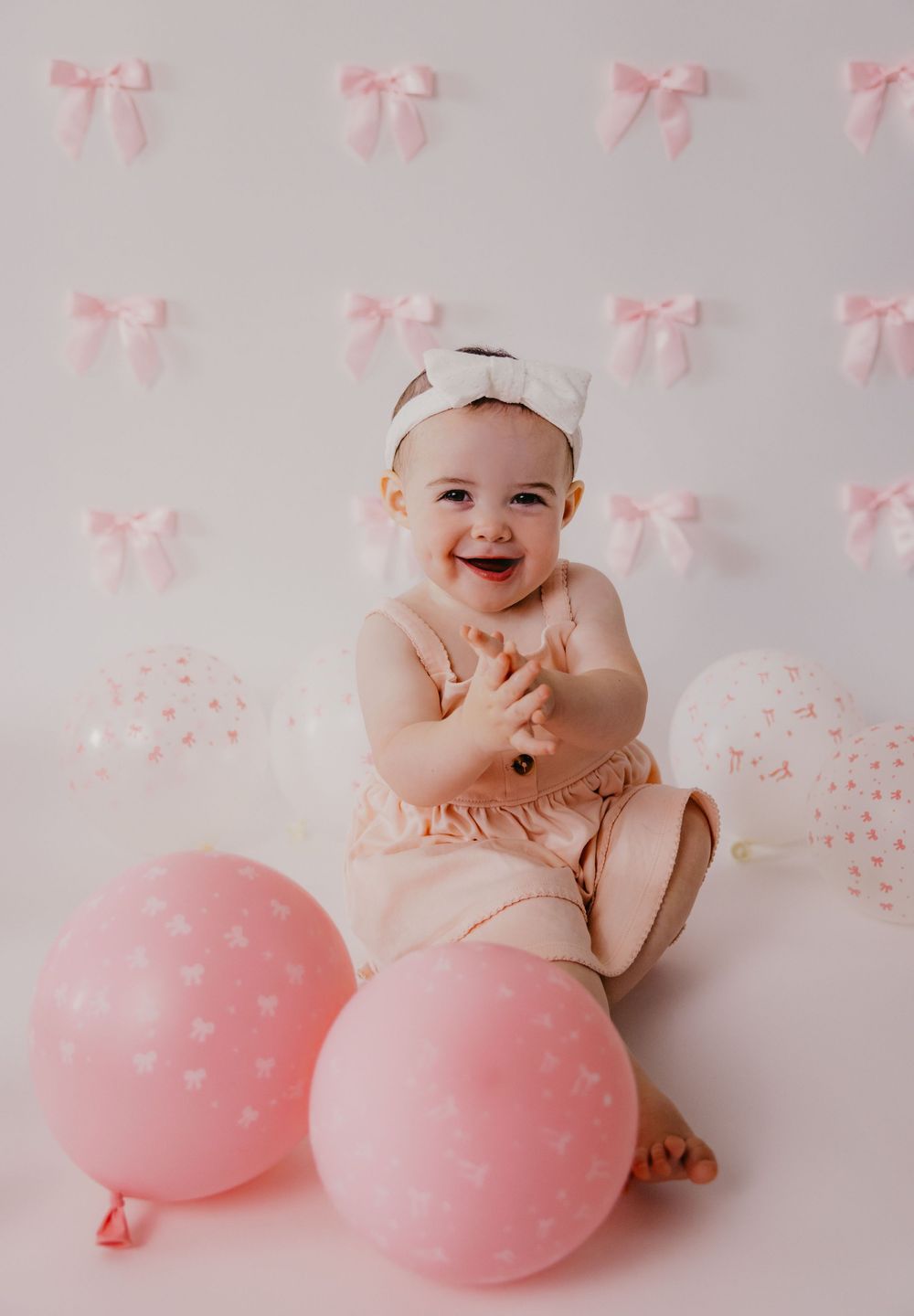 Happy baby girl in pink outfit clapping hands, surrounded by pink balloons and bows backdrop for first birthday cake smash.