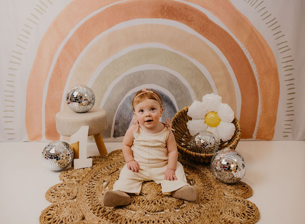 Baby sitting on woven rug with rainbow mural backdrop, disco balls, and daisy decor in a boho-themed photo shoot.