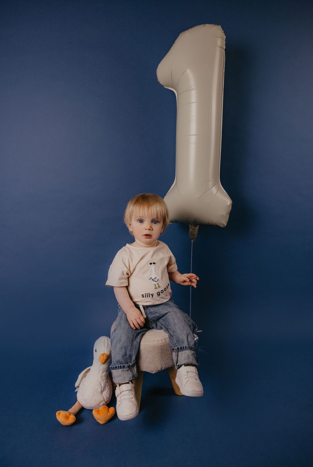 Toddler boy sitting with a number one balloon and a duck toy, celebrating his first birthday against a blue background.