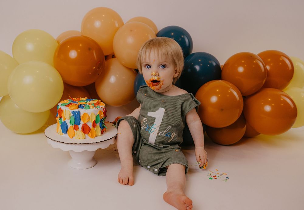 Baby celebrates first birthday with colorful cake and balloon backdrop in olive green romper with number one.