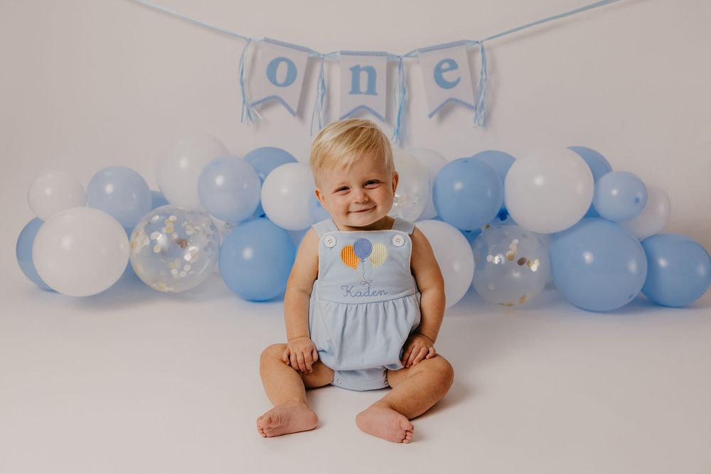 A smiling baby in a blue outfit sits before blue and white balloons and a \"one\" banner for a first birthday.