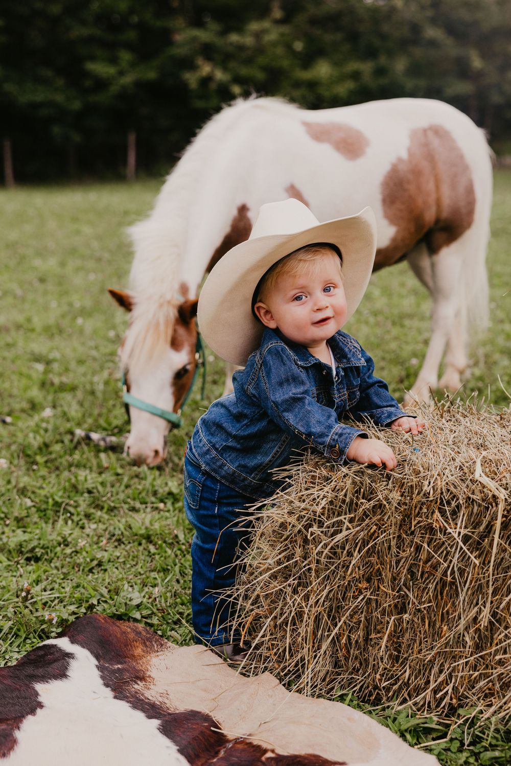 Adorable baby in cowboy hat and denim leans on hay bale beside a paint pony on a green farm.