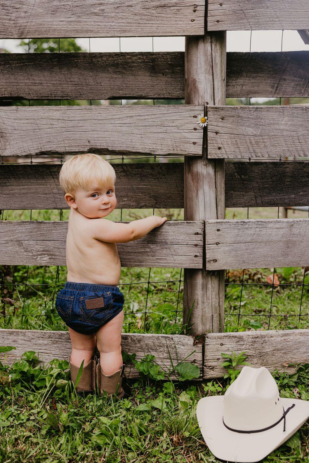 Shirtless toddler in denim shorts and cowboy boots stands by rustic wooden fence with white cowboy hat nearby.