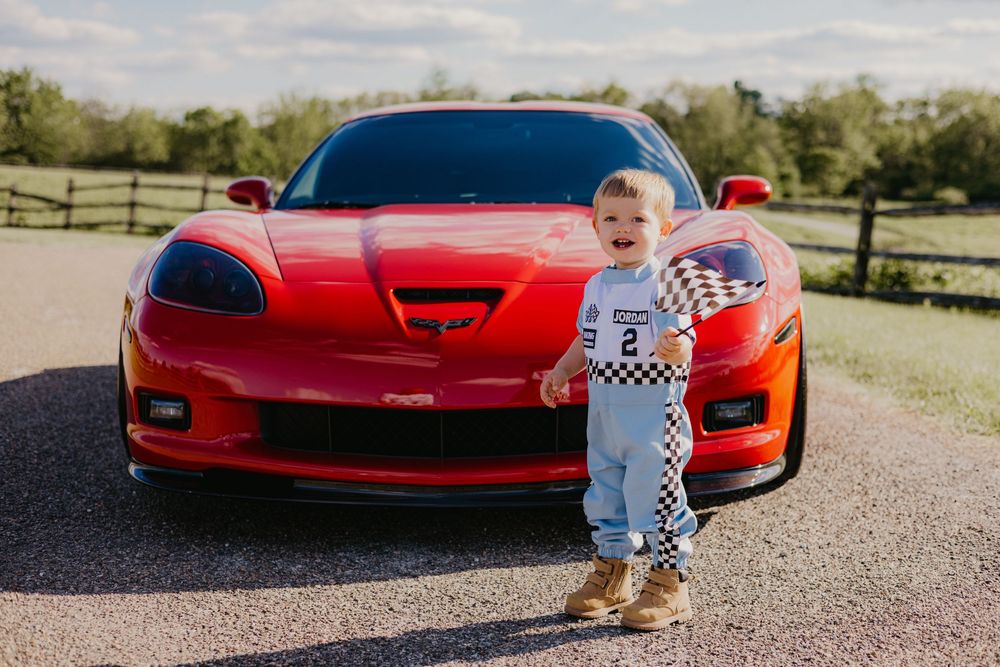 Toddler in racing outfit holding checkered flag in front of red Corvette sports car on a sunny day.