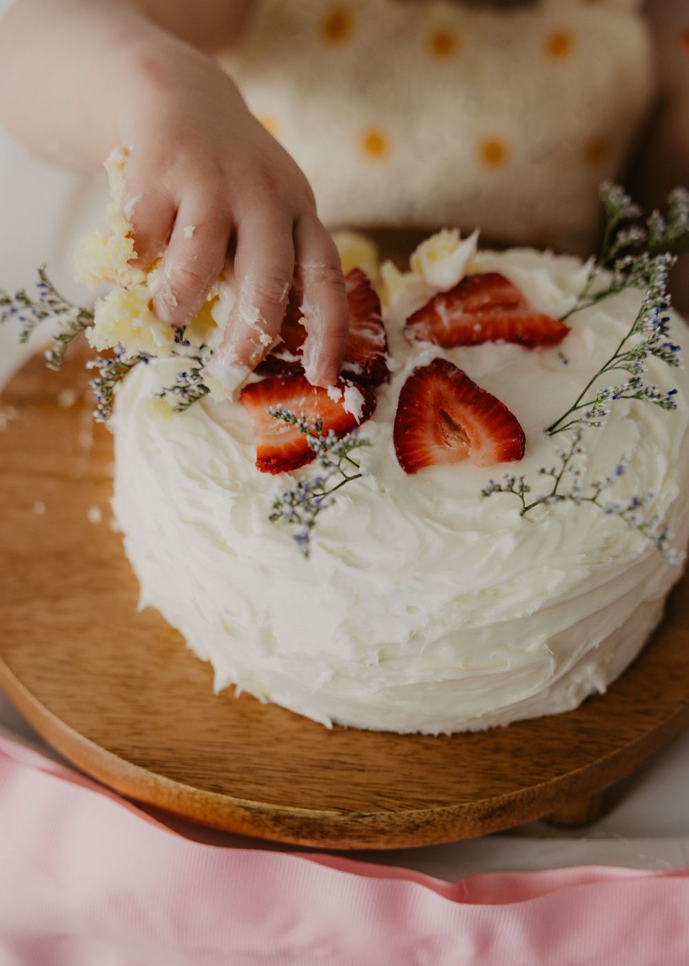 Child's hand reaching into a white frosted cake topped with fresh strawberries and lavender flowers on a wooden board.