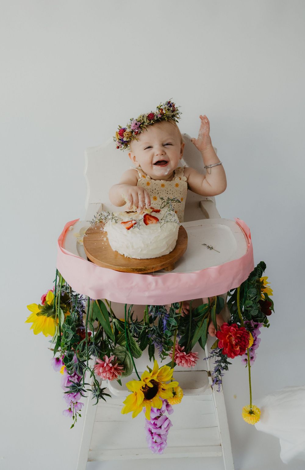 A joyful baby wearing a floral crown smashes a white cake surrounded by colorful flowers during a first birthday cake smash.