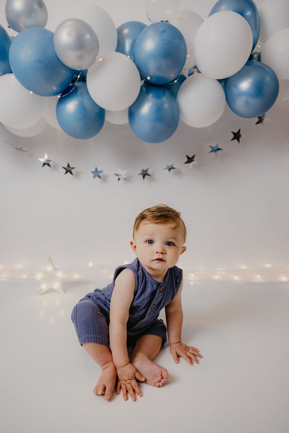 A baby boy in a blue romper sits on a white floor surrounded by blue and white balloons and star decorations.