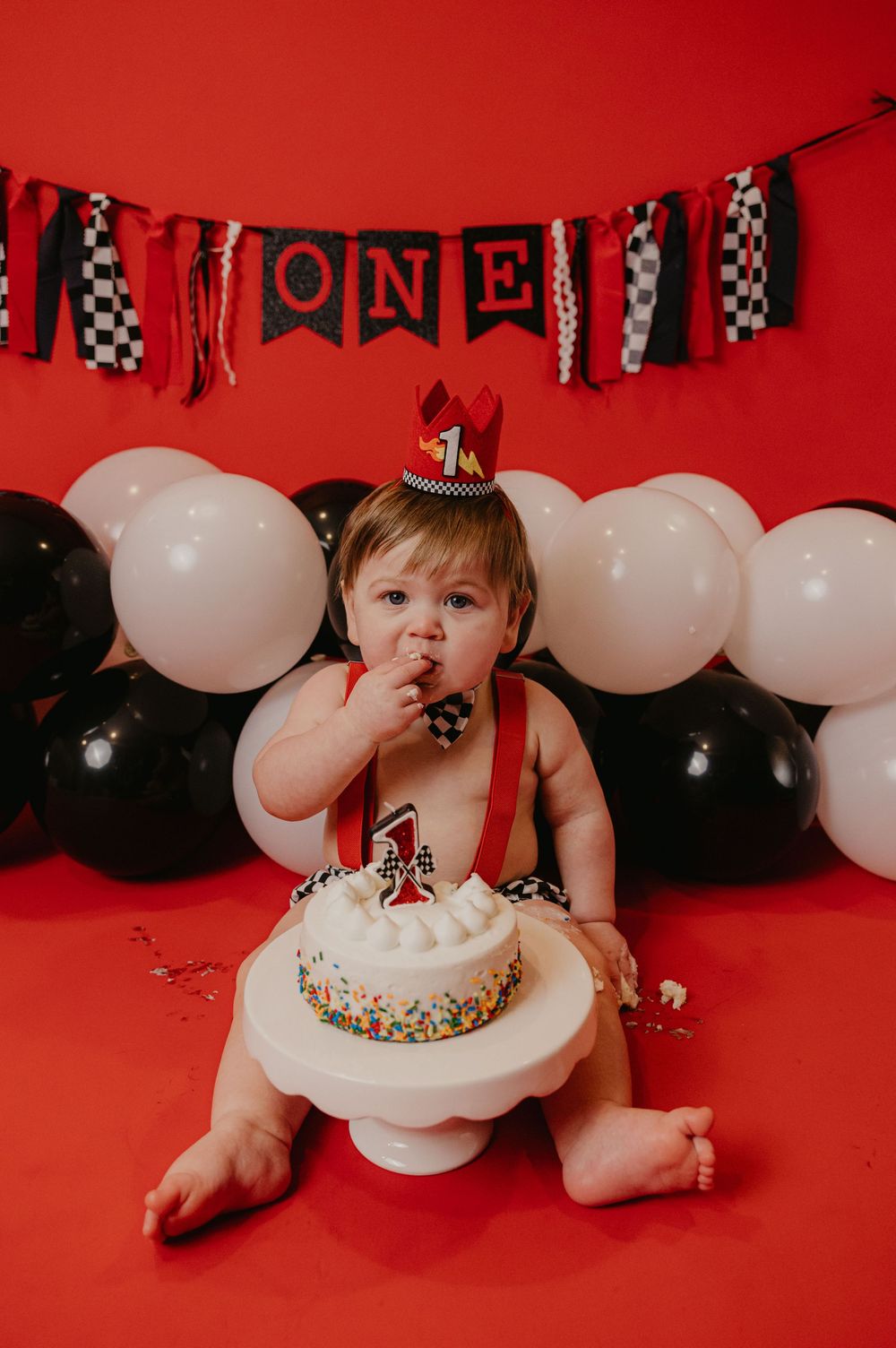 Baby's first birthday cake smash on red background with ONE banner, black and white balloons, wearing racing theme outfit.