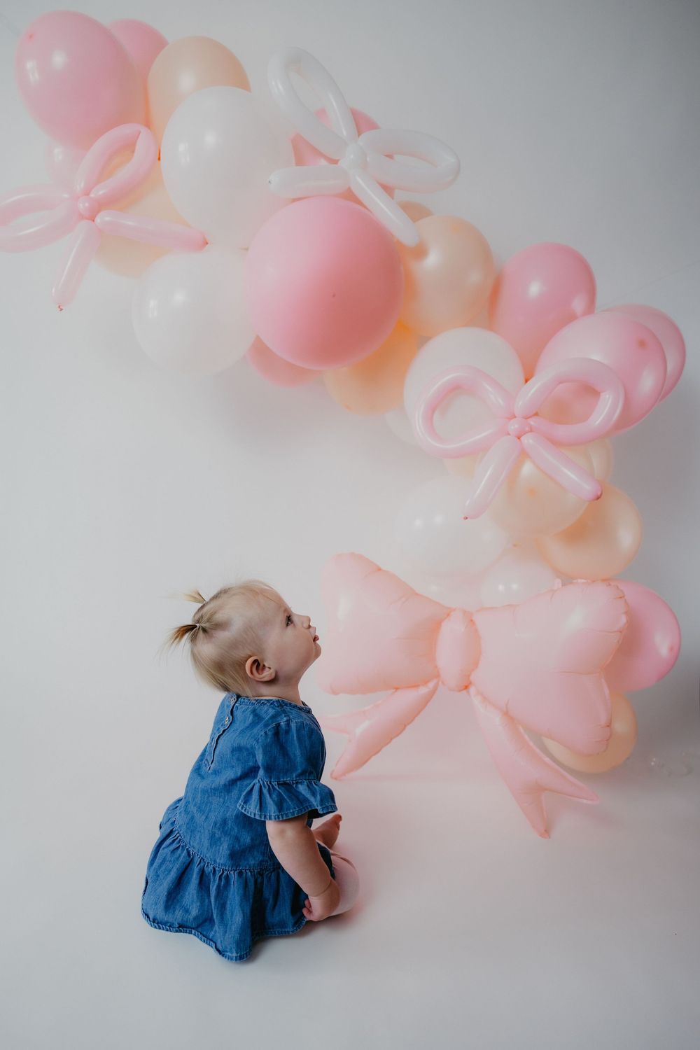 A baby girl in a blue dress sits on a white floor gazing up at pink and white balloon decorations with bow shapes.