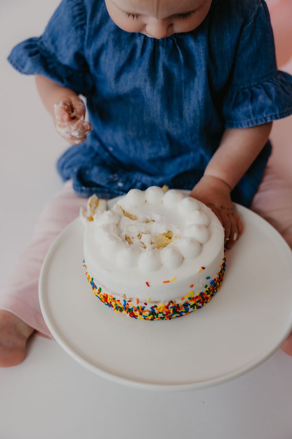Baby girl in blue dress touching white birthday smash cake with colorful sprinkles on white plate.