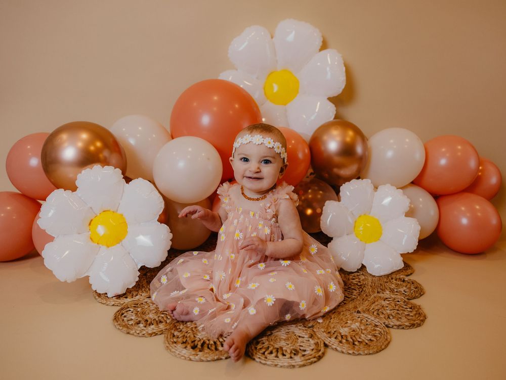 Baby girl in pink dress sits among daisy balloons and rose gold balloons for a first birthday cake smash photo.