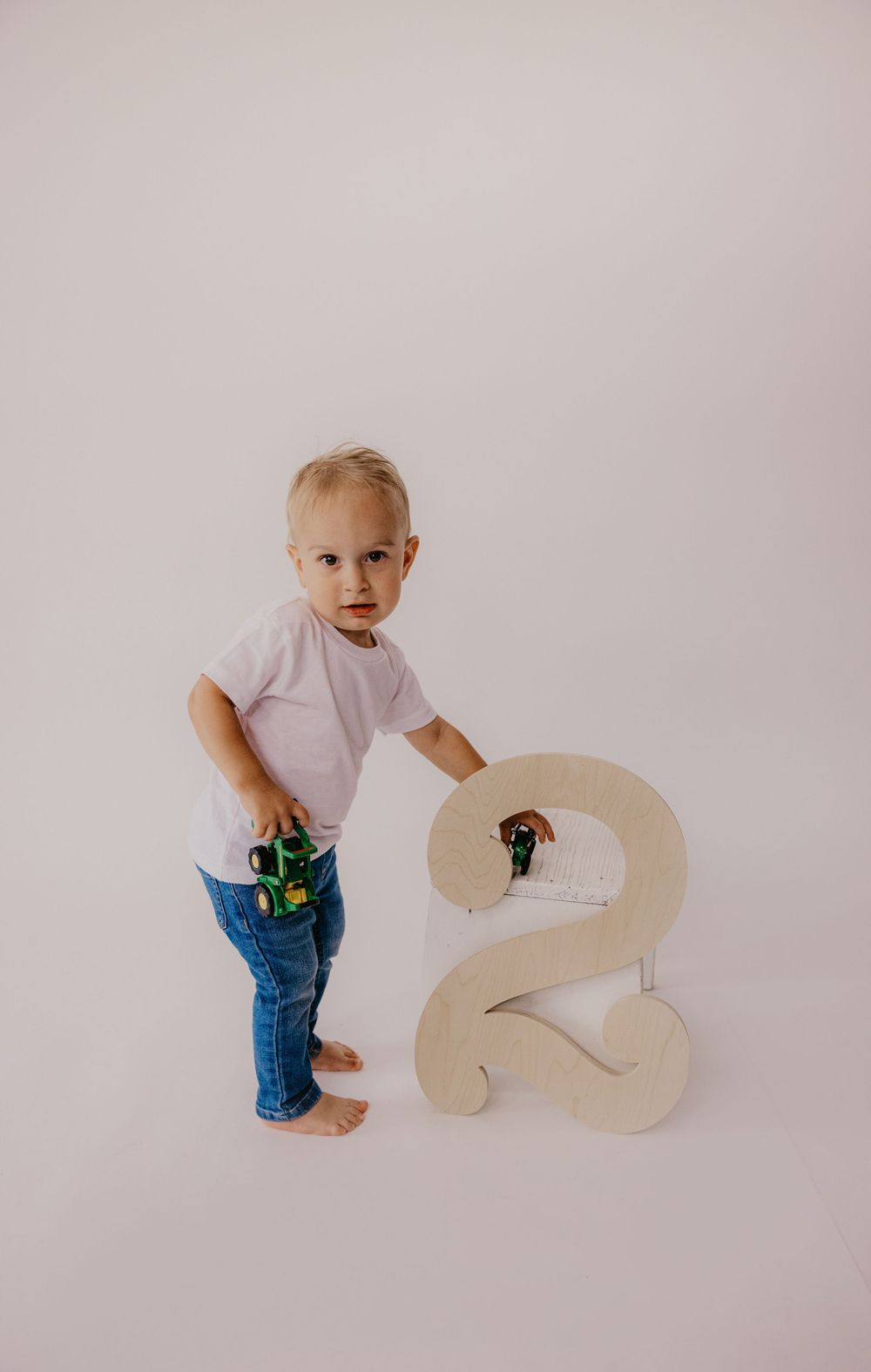 Toddler boy in white tee and jeans holds toy tractor beside large wooden number 2 for second birthday photo.