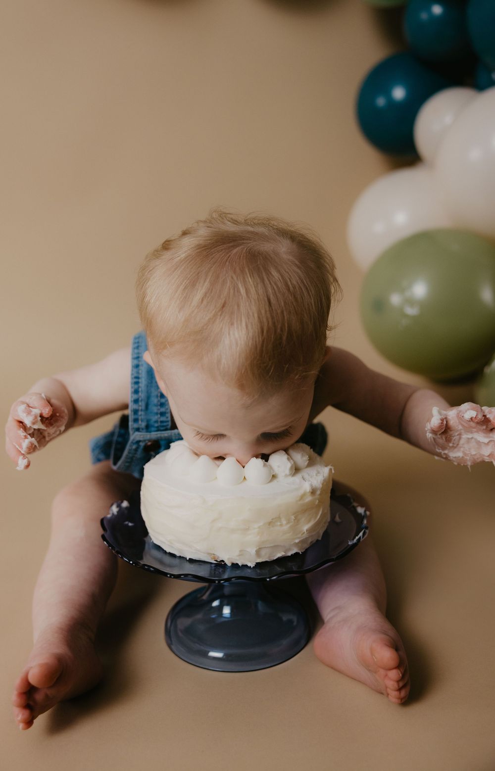 Baby in denim romper smashing face into white frosted first birthday cake on black stand with colorful balloons.