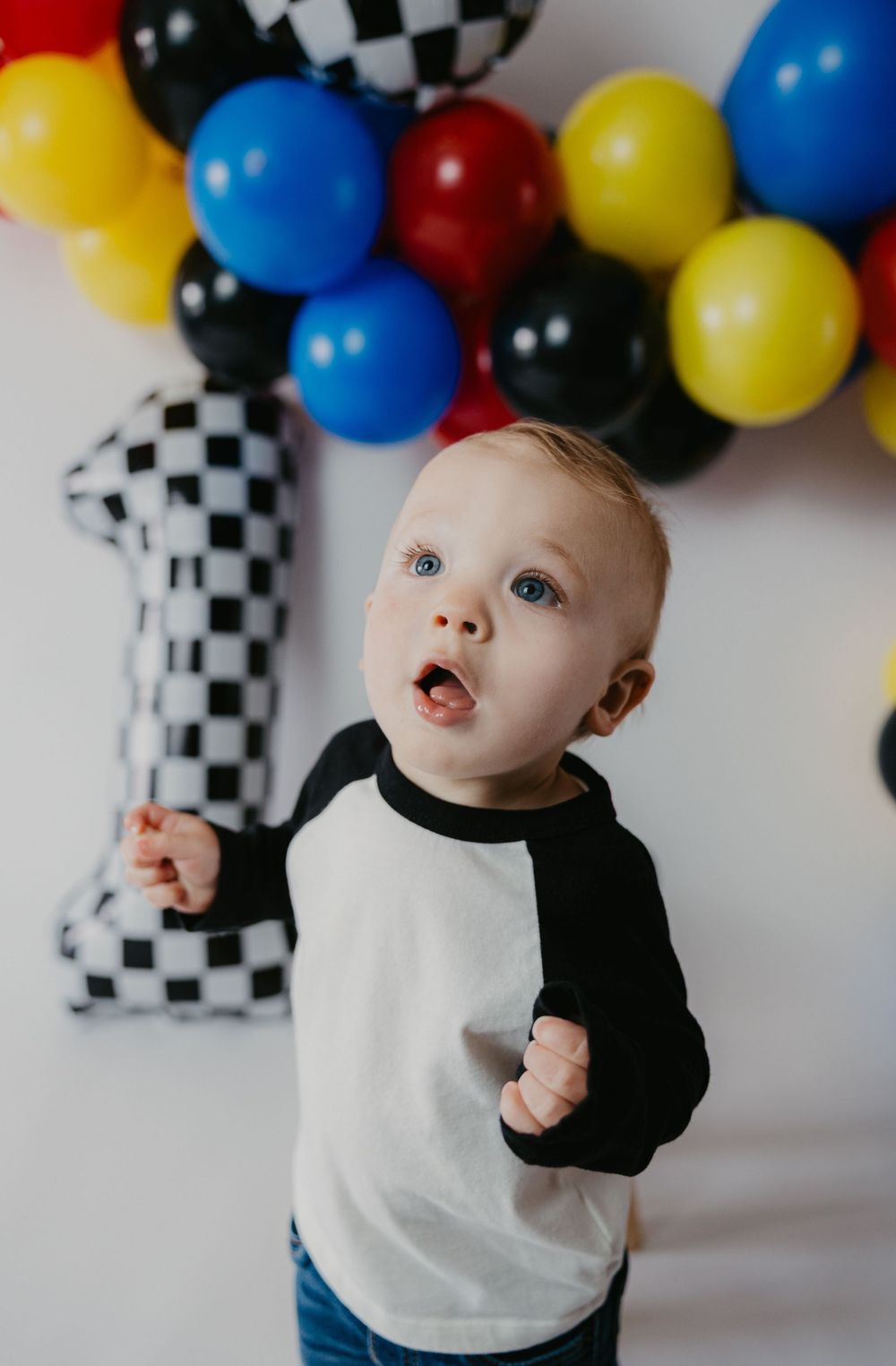 Toddler boy in raglan shirt at race car themed first birthday party with checkered number one balloon.
