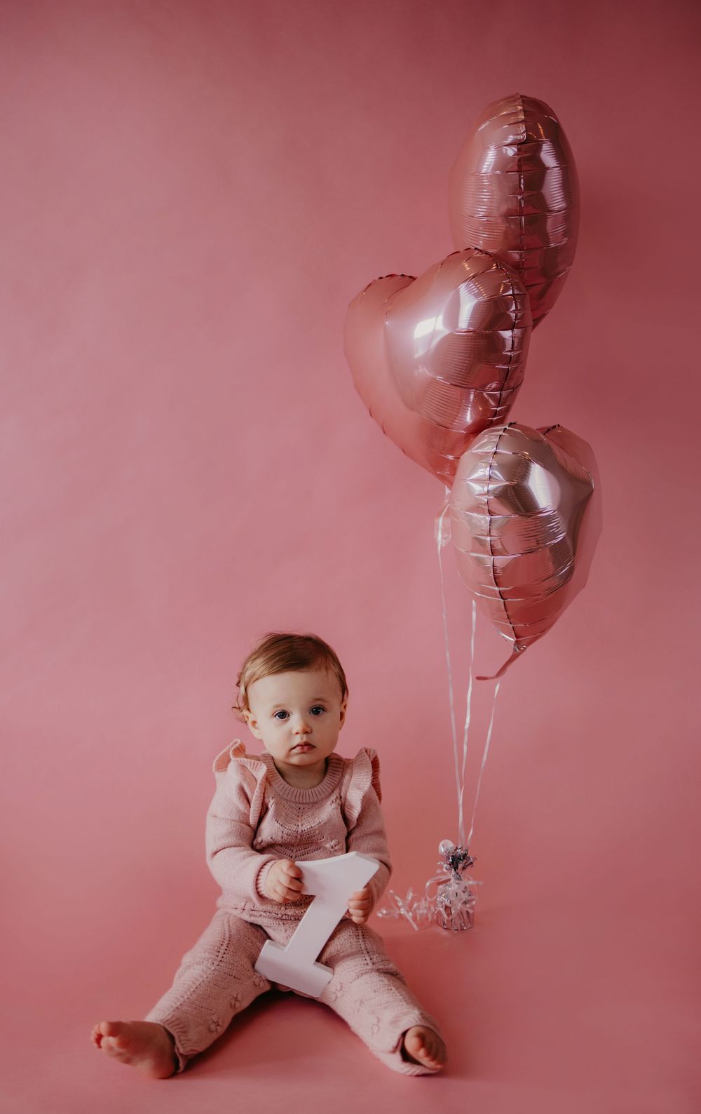 Baby girl in pink knit outfit sits beside heart-shaped pink foil balloons against a pink background.