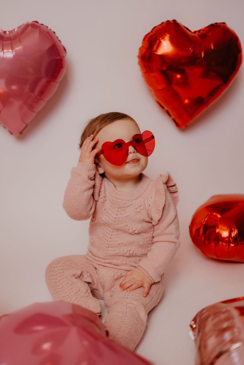 Cute toddler in pink knit outfit wearing red heart sunglasses surrounded by pink and red heart-shaped balloons.