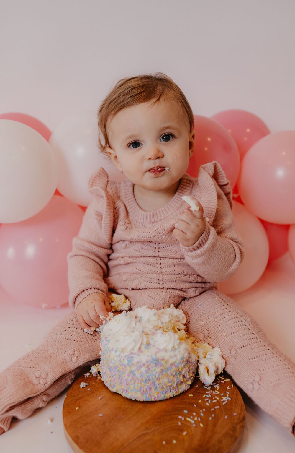 Baby girl in pink knit outfit smashes first birthday cake surrounded by pink balloons.