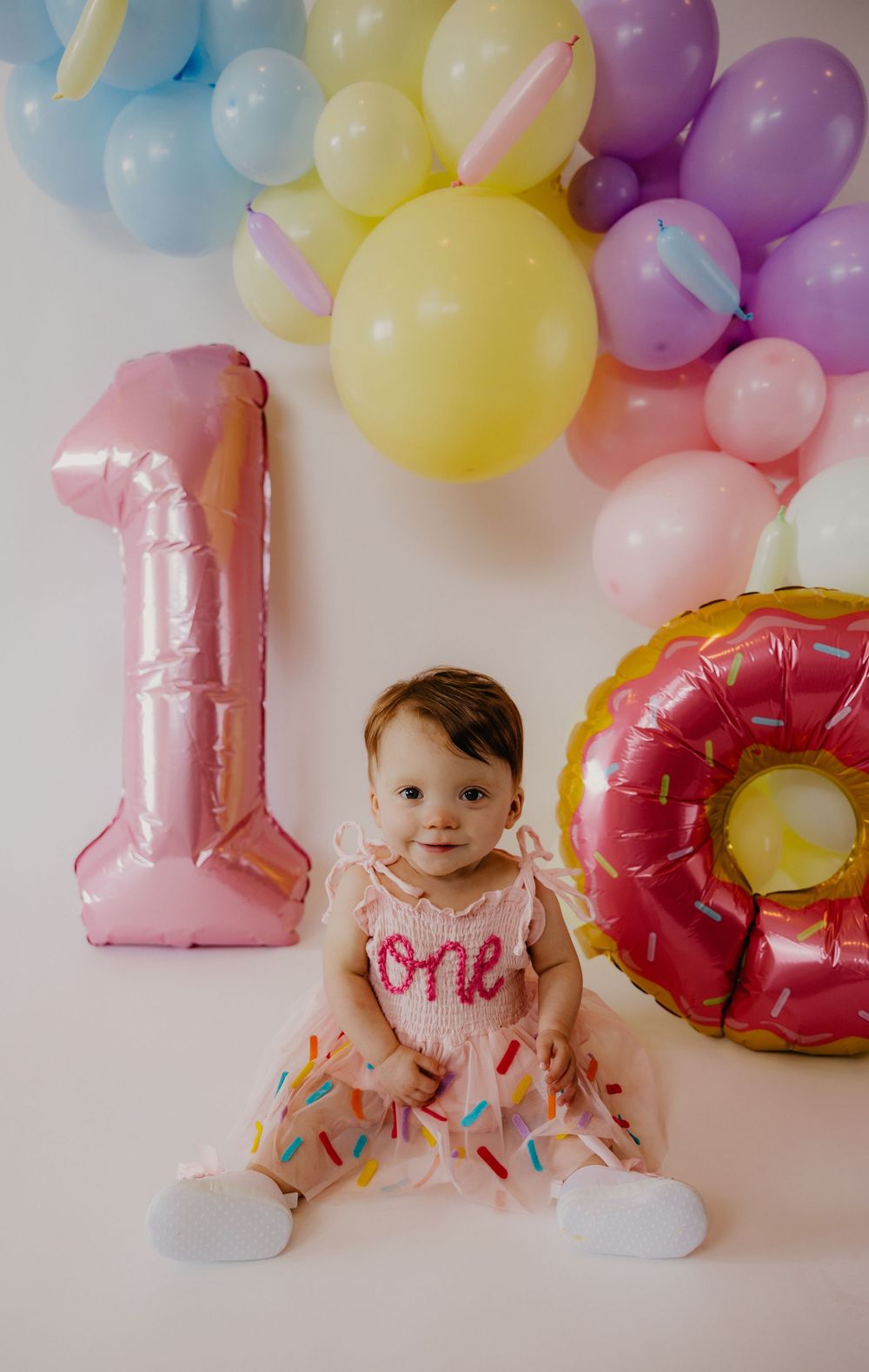 Baby girl in pink donut dress sits between number 1 balloons surrounded by pastel balloon garland for first birthday.