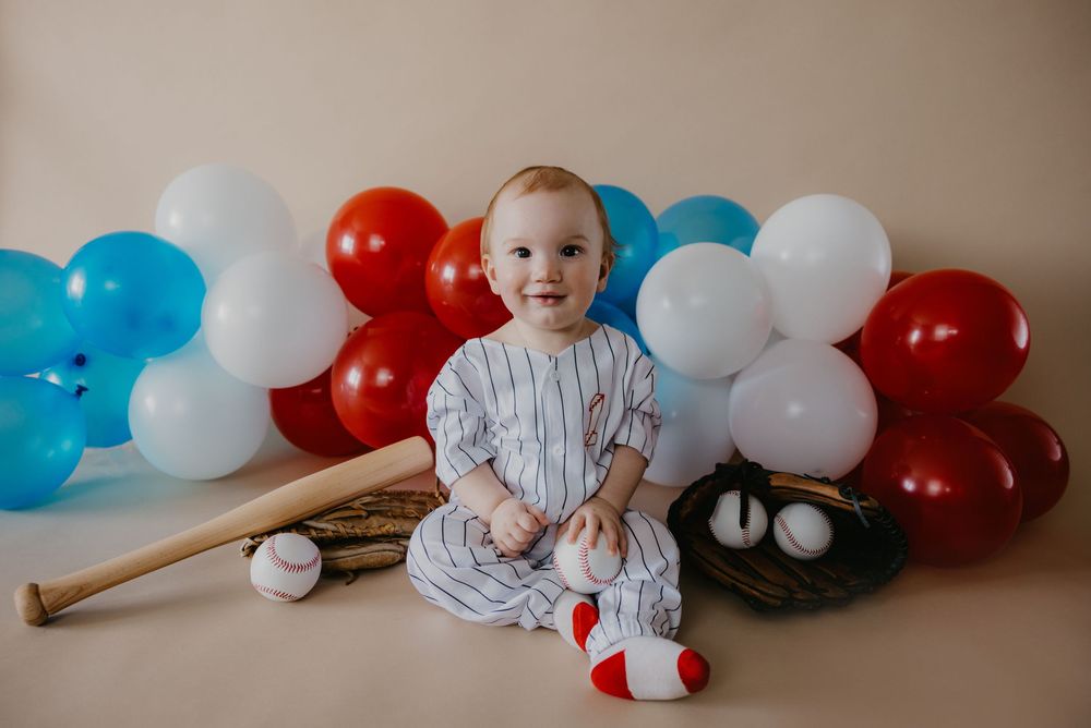 Baby in baseball outfit sits with bat, glove, and red, white, and blue balloons for a sports-themed photo.