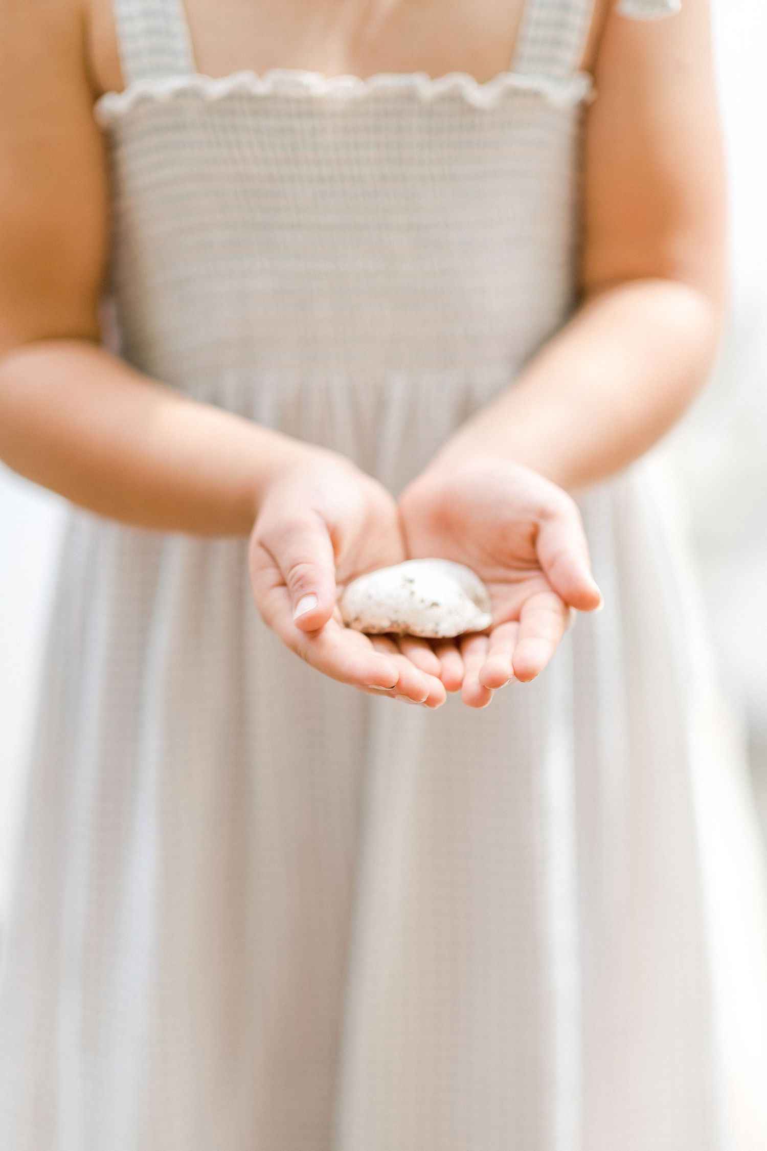 Young girl in smocked dress holding a small white seashell in her cupped hands.