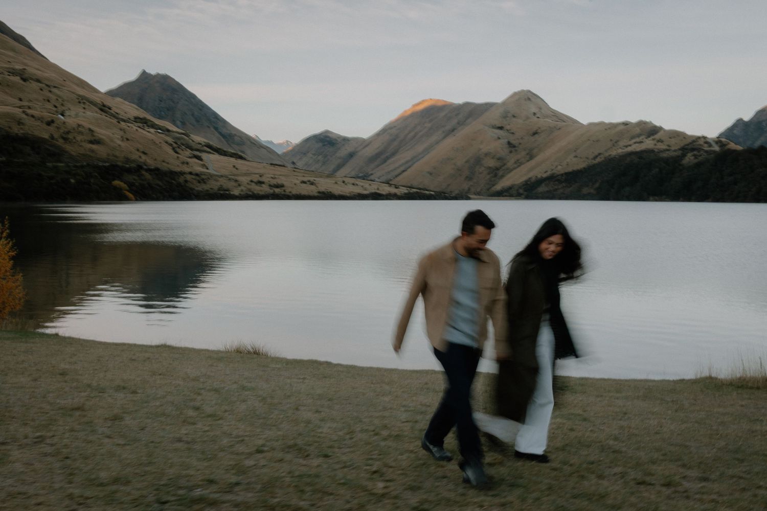 Couple walking hand in hand by a serene mountain lake at dusk in New Zealand.
