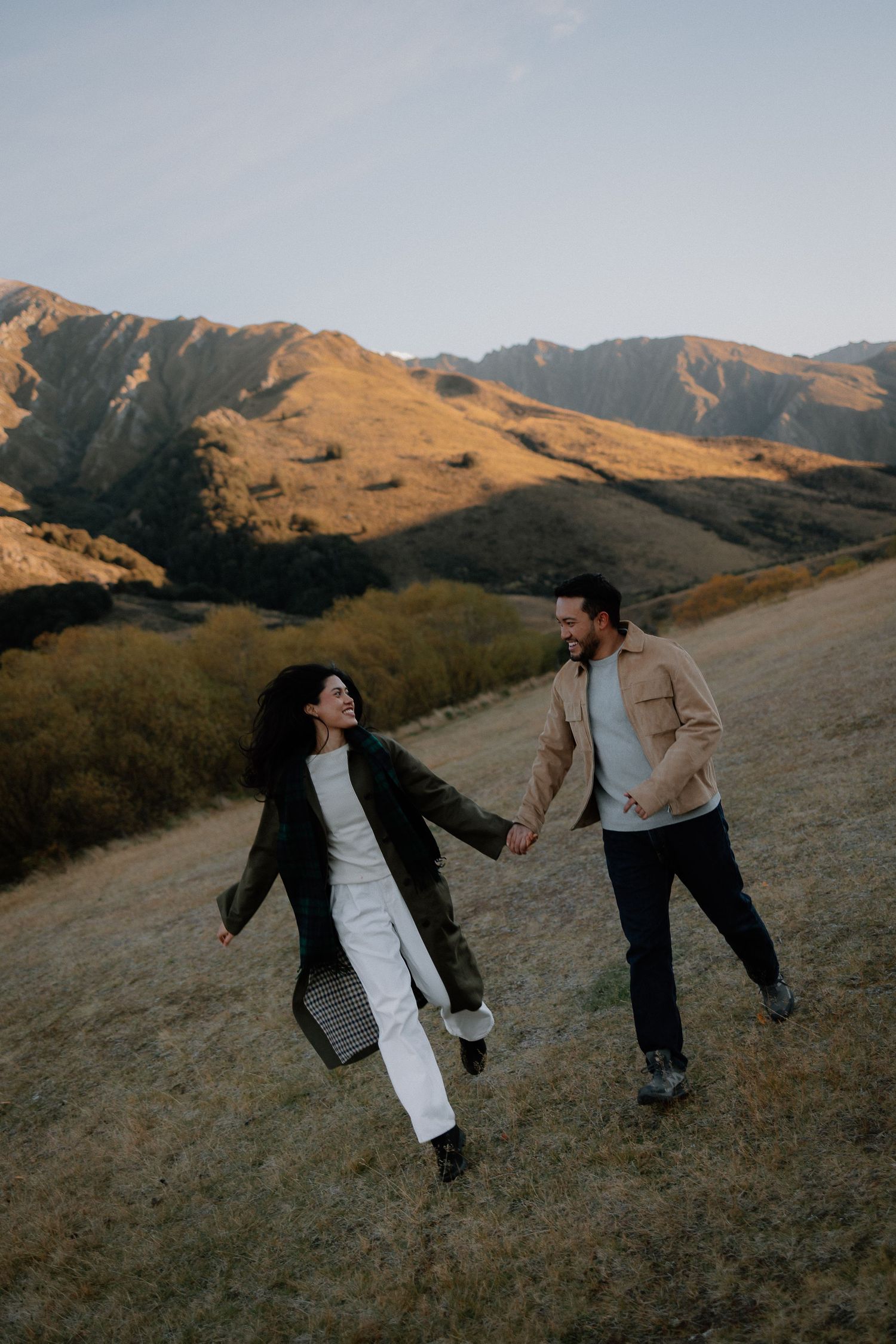 A joyful couple holds hands while walking through a scenic mountain valley landscape at golden hour.
