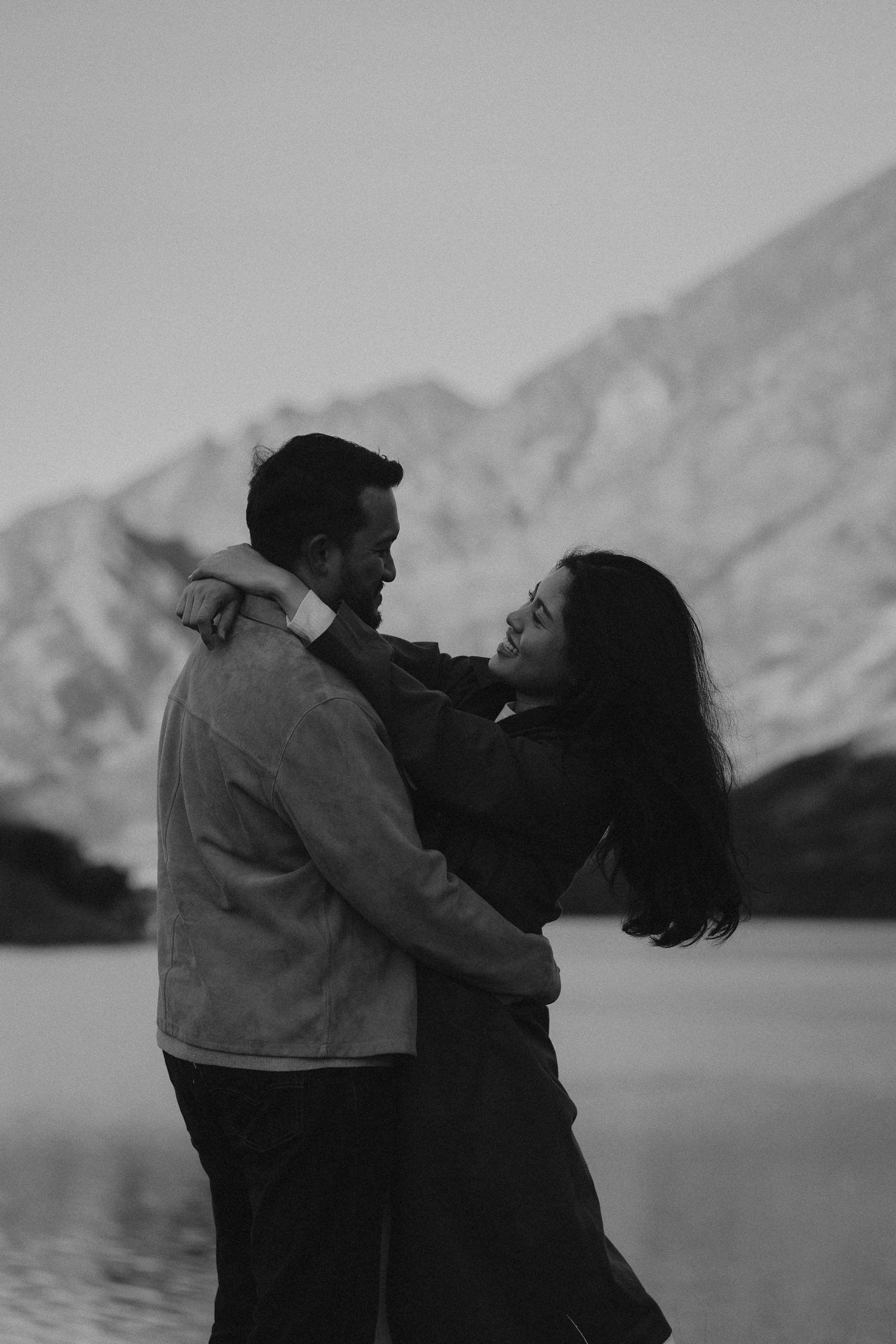 Black and white photo of a couple embracing and smiling near a mountain lake at dusk.