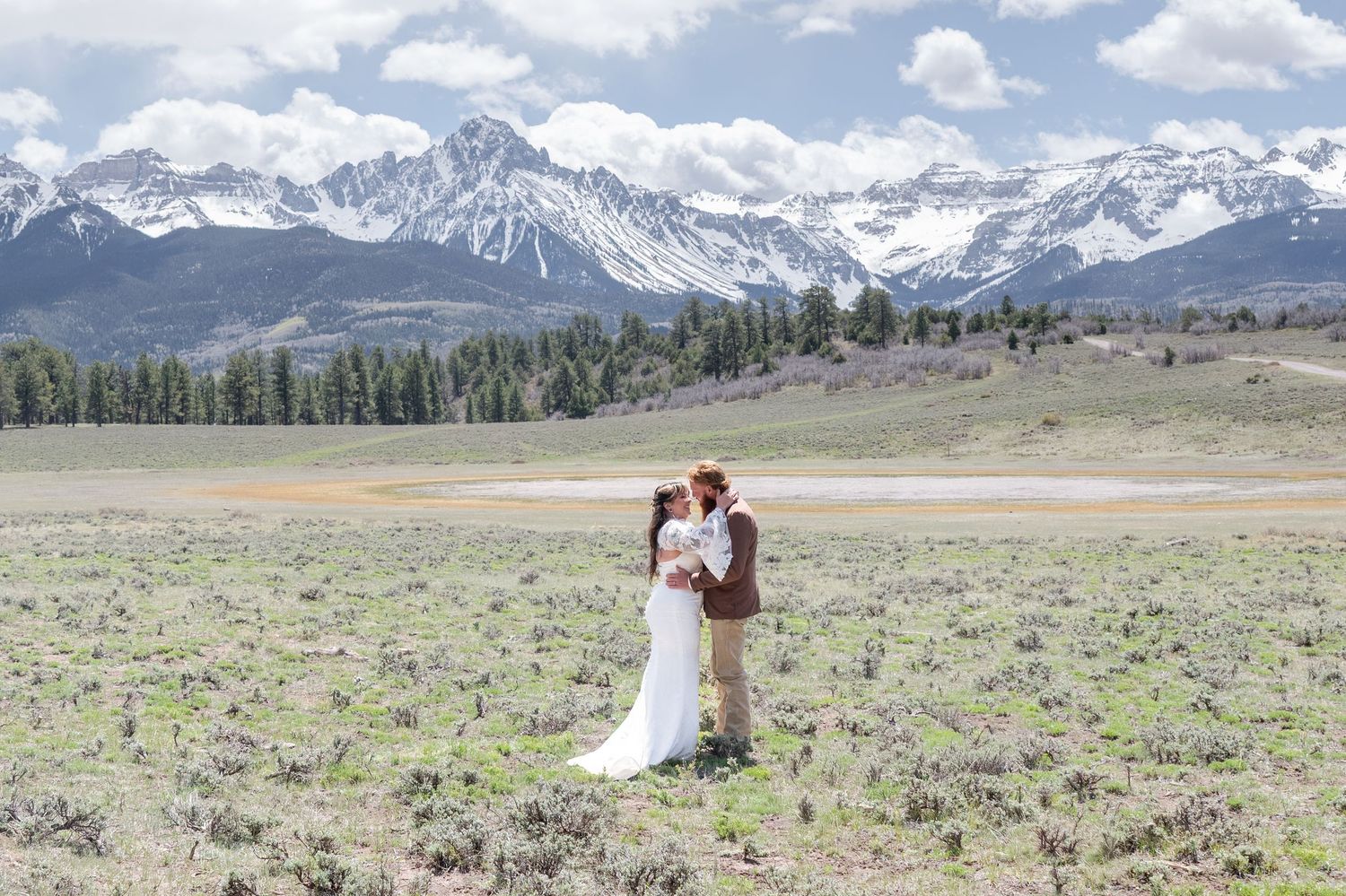 A bride and groom stand in an intimate embrace in a large open meadow. The San Juan mountains are in the background with snow on the peaks.