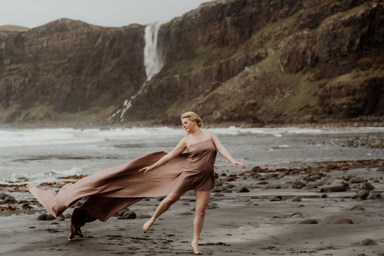 Woman in flowing brown dress dancing on rocky beach with dramatic waterfall and green cliffs in background.