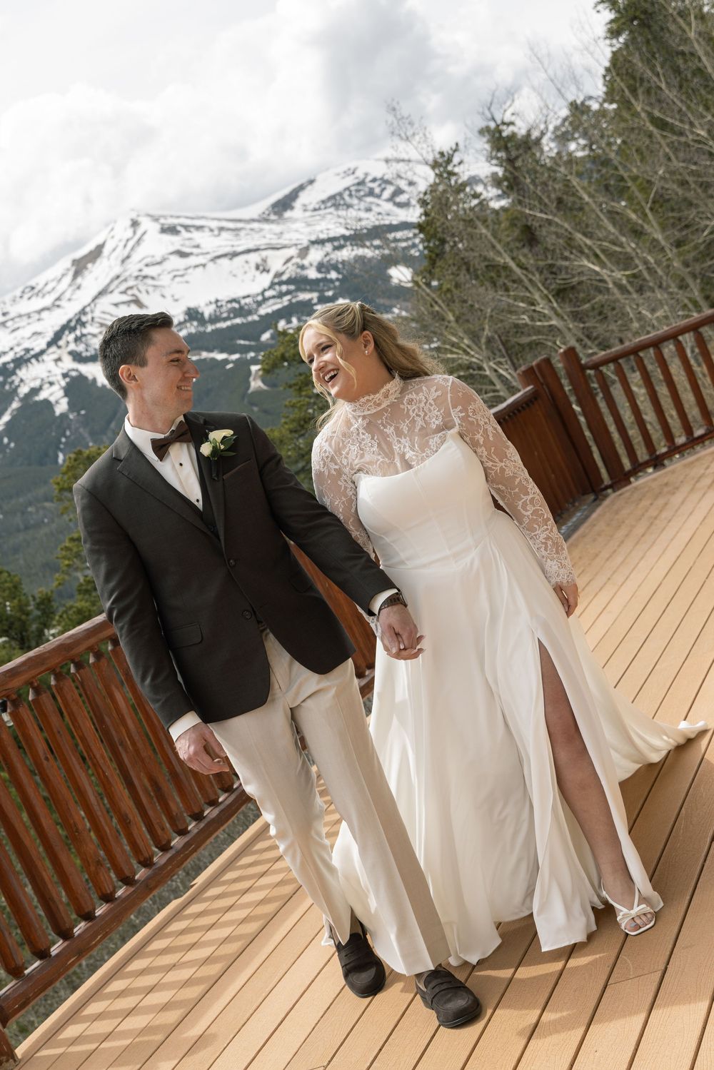 A bride and groom walk hand-in-hand on a wooden deck with snow-capped mountains in the background.