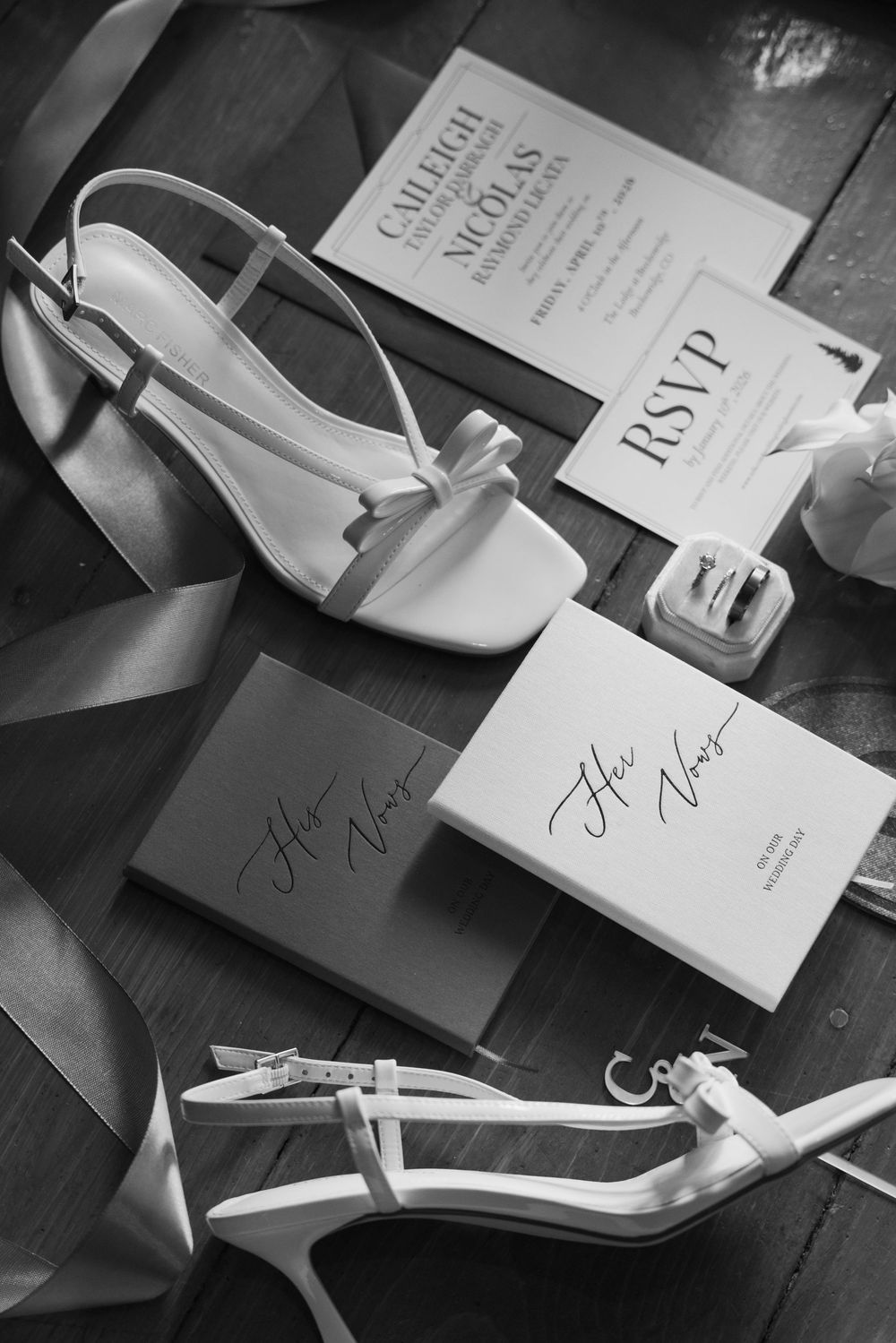 Black and white flat lay of bridal heels, wedding invitations, RSVP card, and vow booklets on wood.