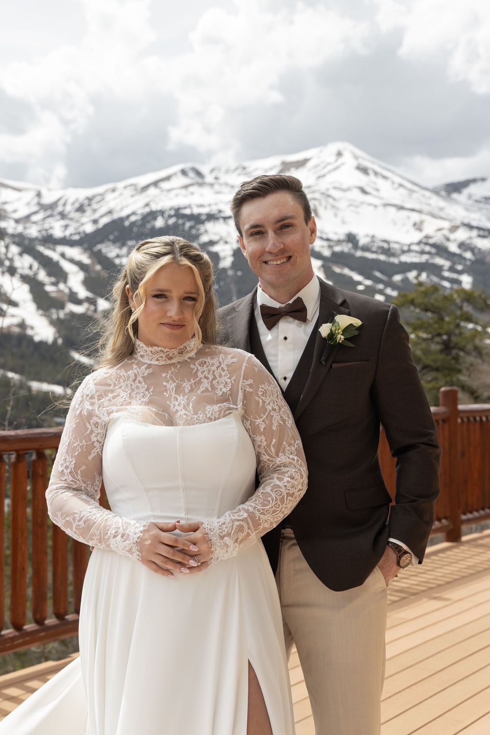 Bride and groom pose on snowy mountain deck, she wears lace gown, he wears brown tuxedo with boutonniere.