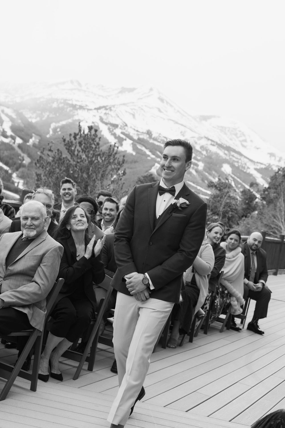 Groom smiling at wedding ceremony on mountain deck with snowy peaks backdrop, guests applauding in black and white photo.