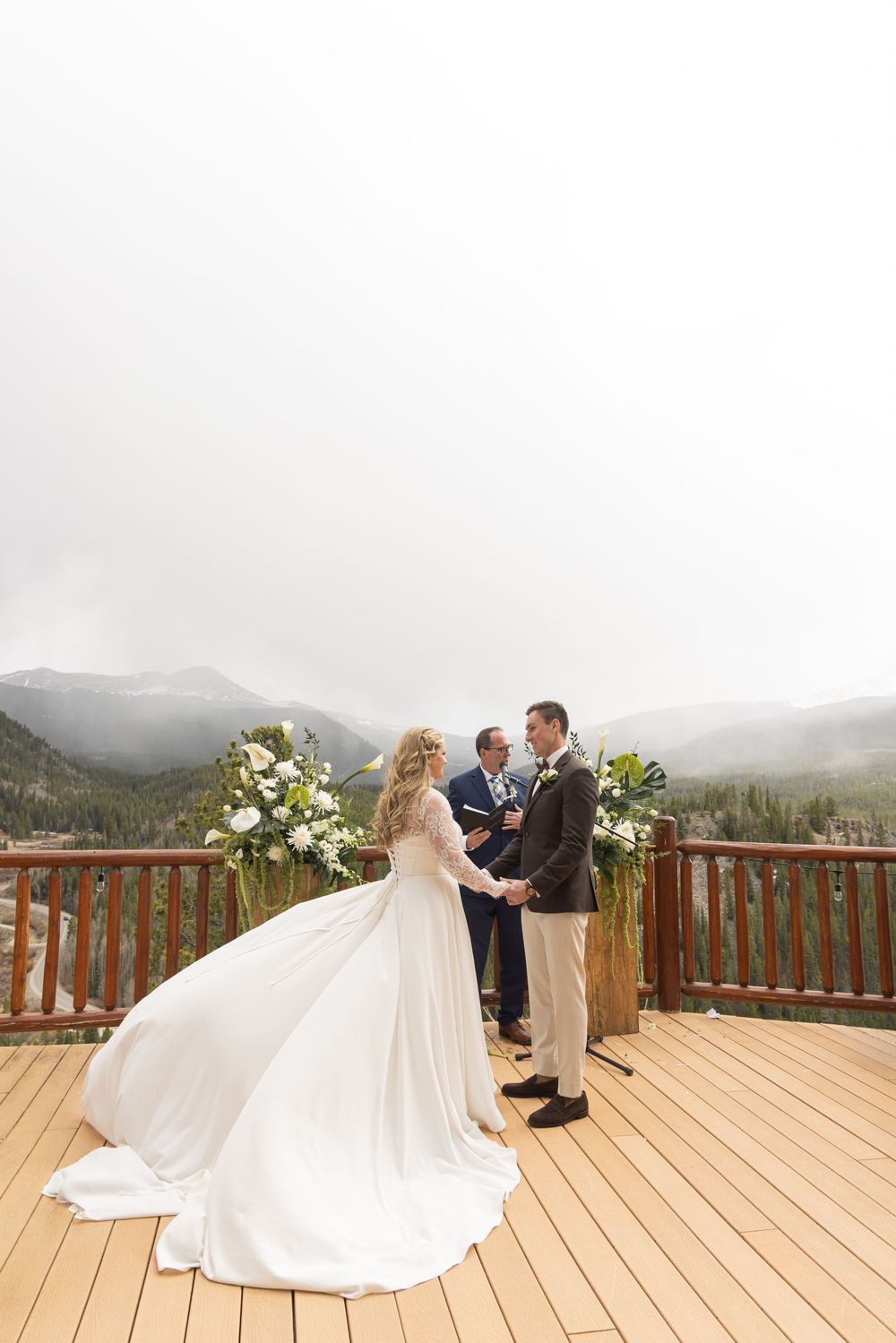 Bride and groom exchange vows on a mountain deck with misty peaks and white floral arrangements at an outdoor wedding ceremony.