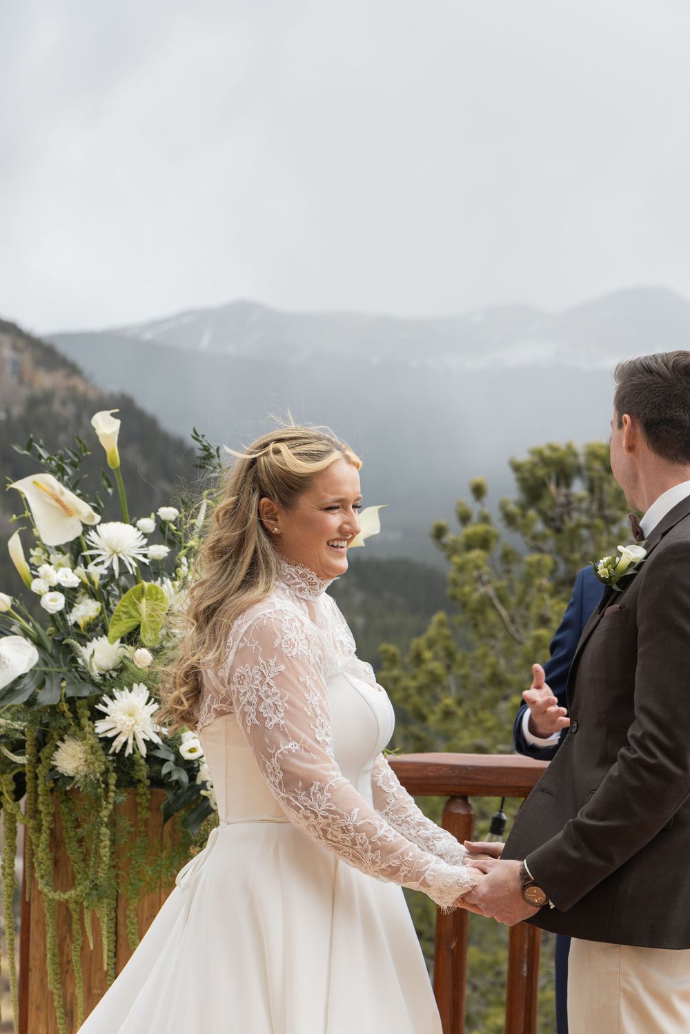 Bride smiling joyfully at groom during outdoor mountain wedding ceremony with white floral arrangements.