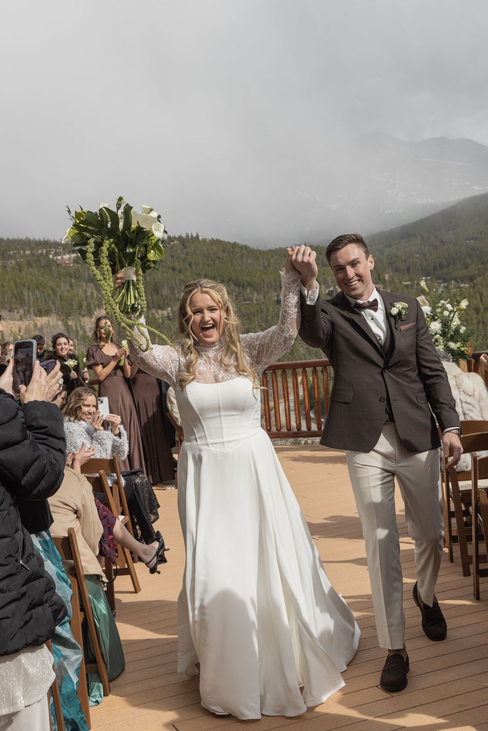 Joyful newlyweds celebrate their outdoor mountain wedding ceremony, bride raising bouquet, groom in gray suit, guests seated.