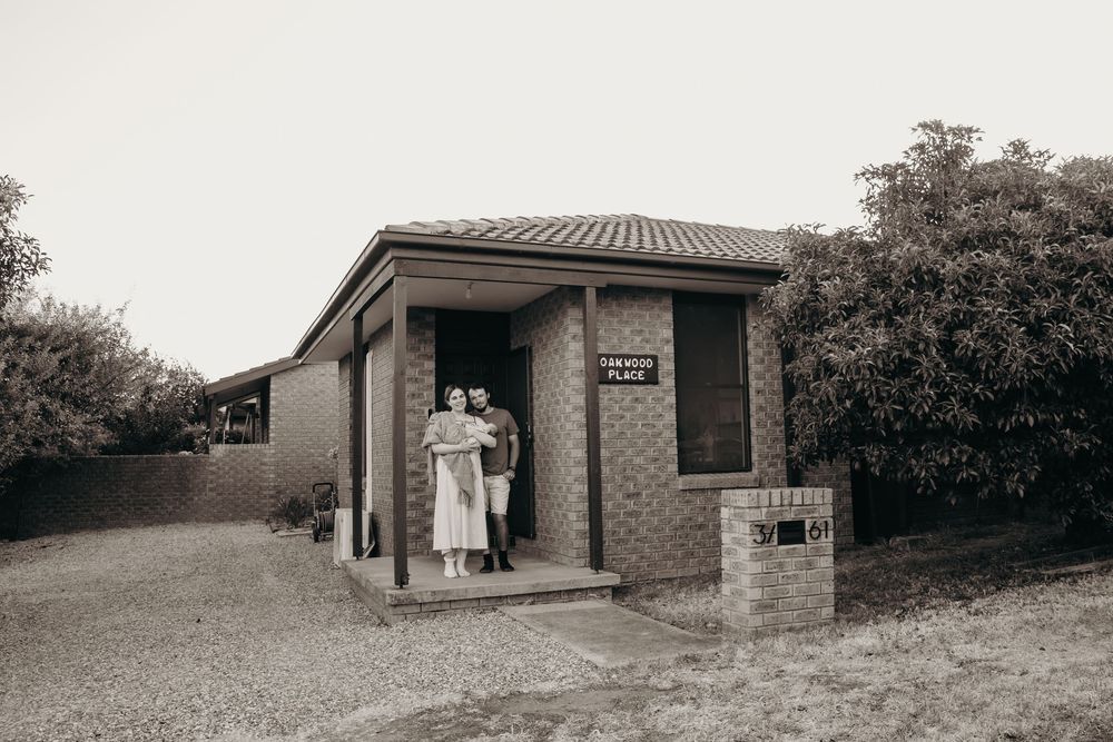 A vintage black and white photo of a couple standing outside a small brick home with a letterbox.