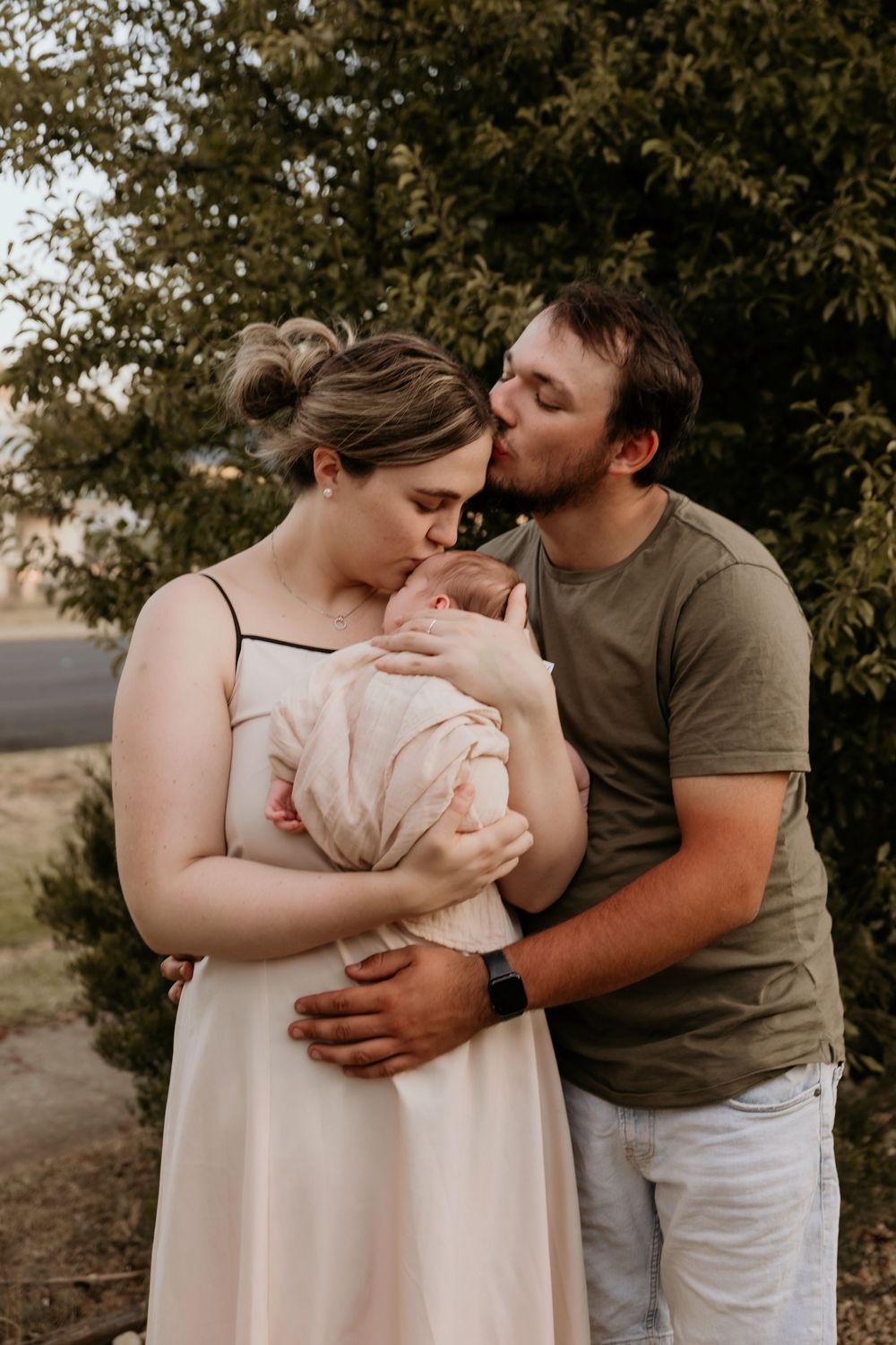 Parents kissing their newborn baby together in an outdoor setting.