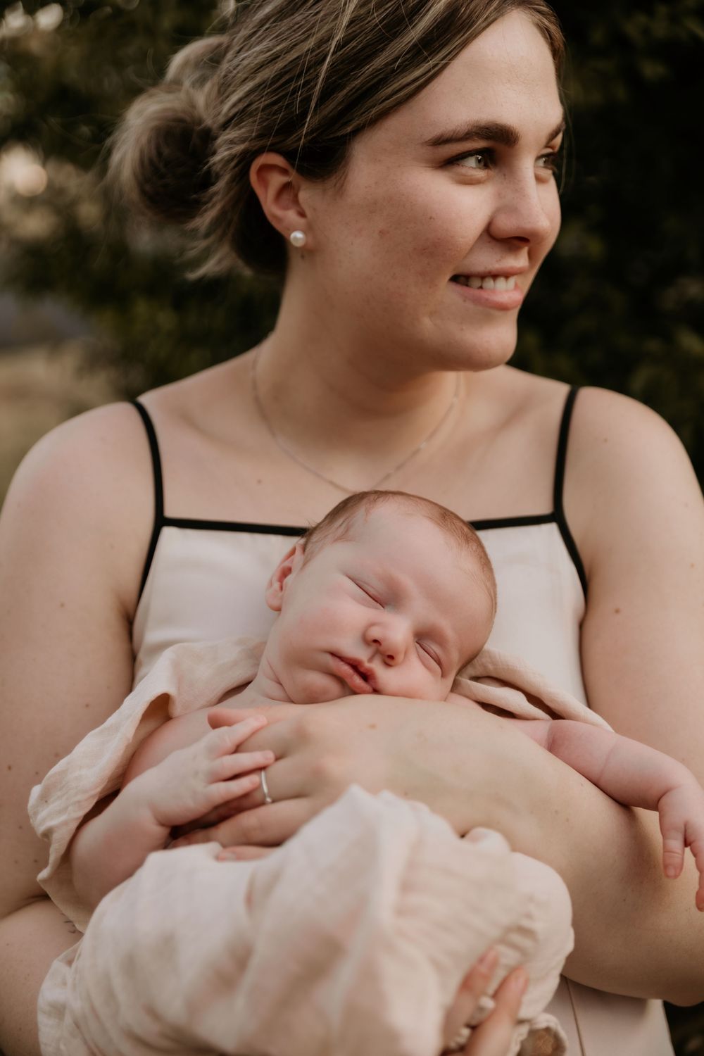 Smiling mother holding sleeping newborn baby against her chest outdoors.