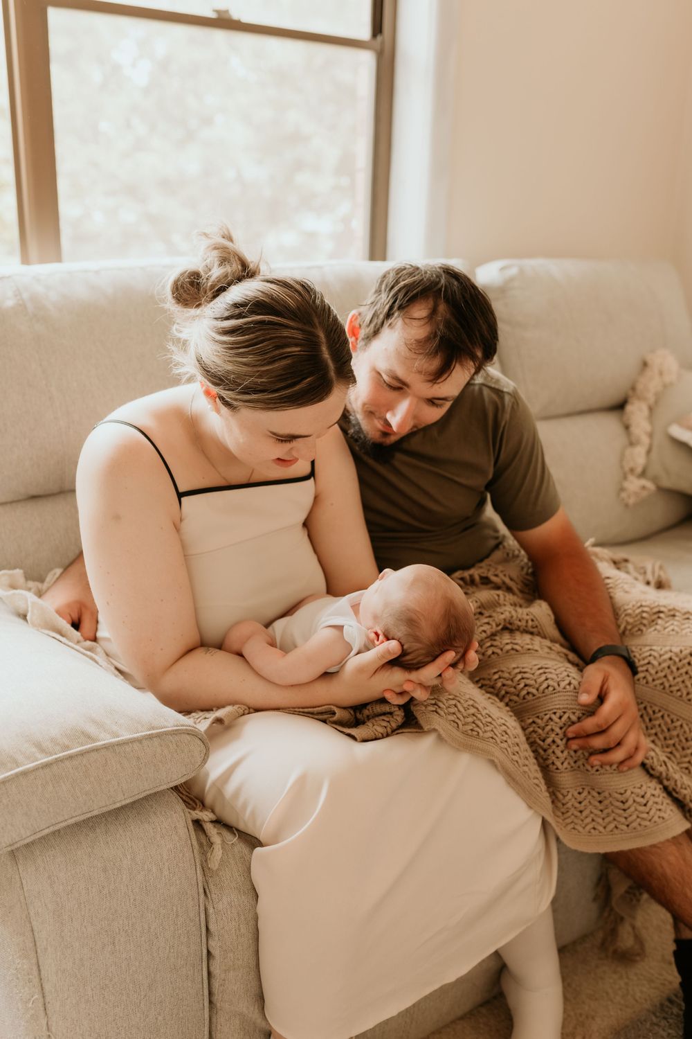 New parents sitting on a couch together lovingly gazing down at their newborn baby in warm light.