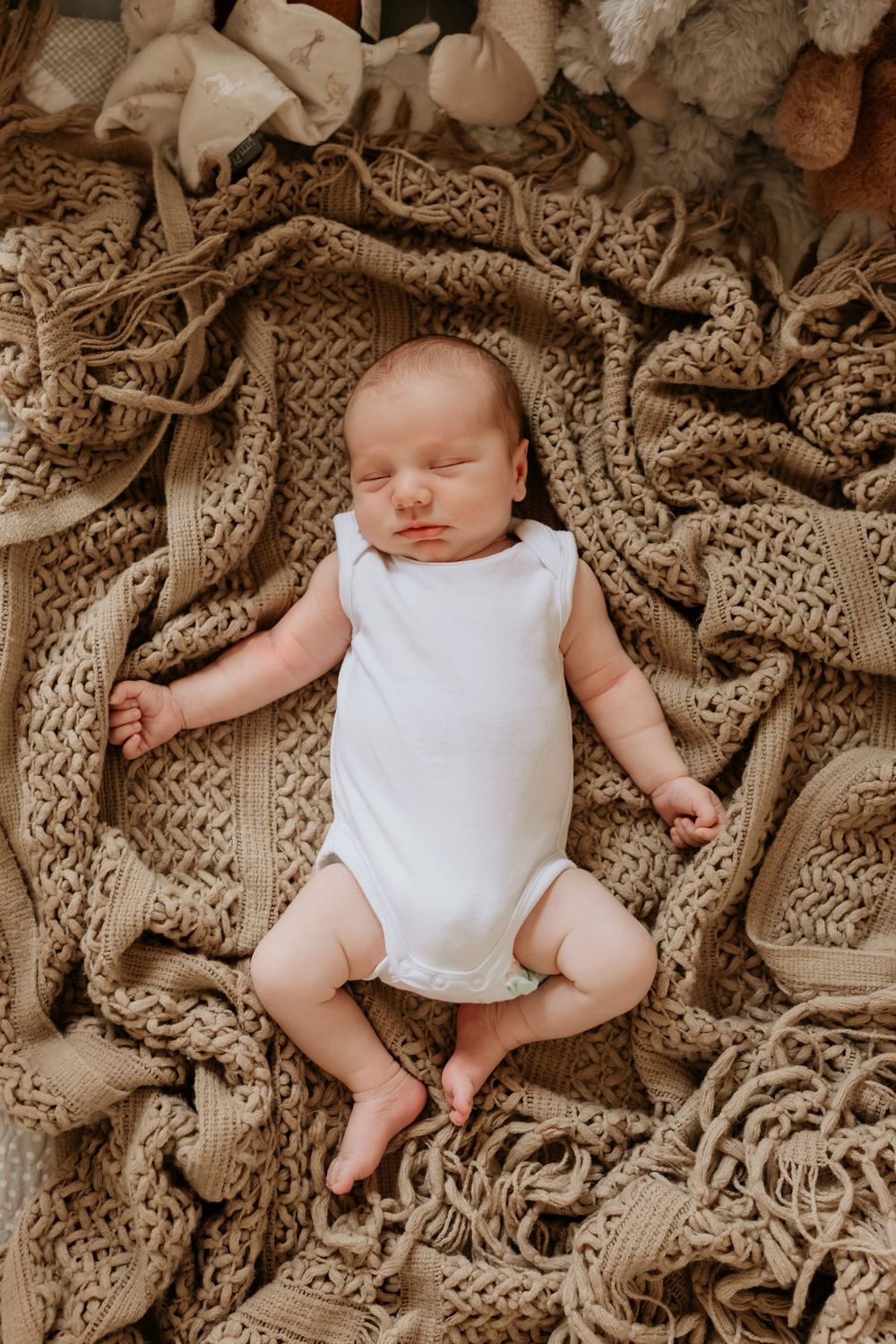 Peaceful newborn baby sleeping in white onesie on chunky knit beige blanket.