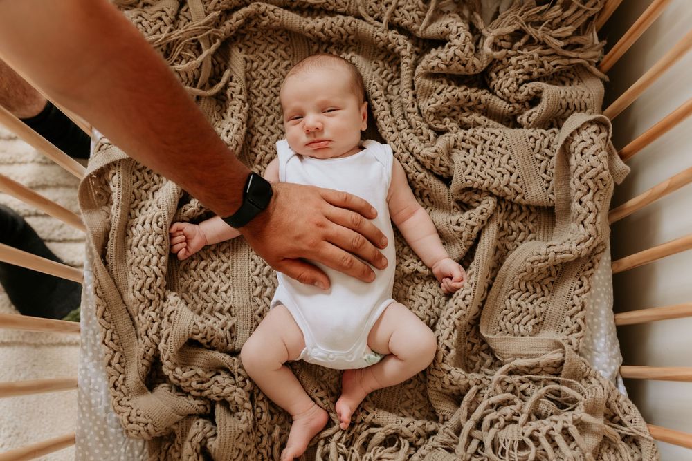 Father gently placing hand on sleeping newborn baby lying in crib on knit blanket.