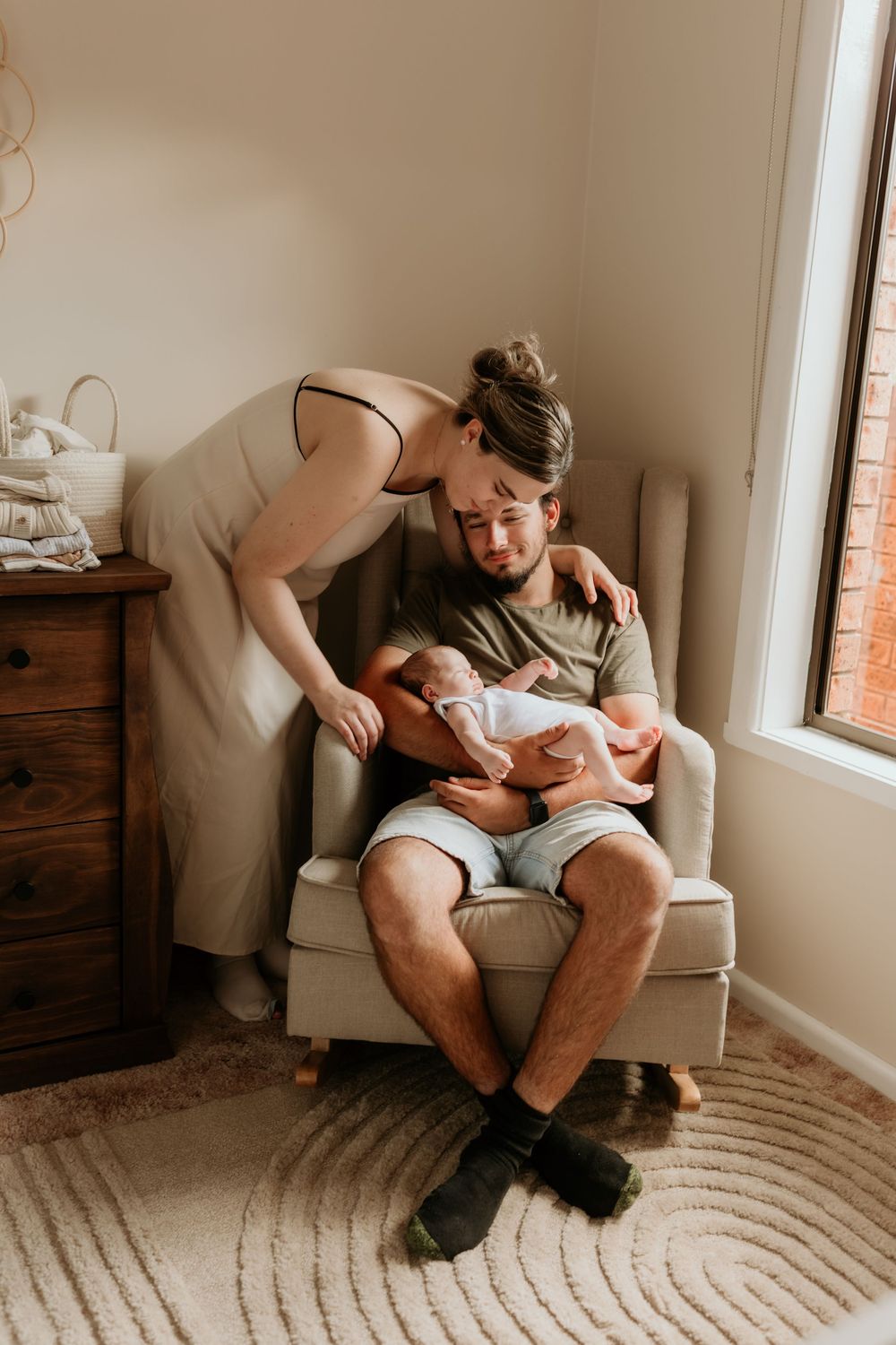 Mother kissing father on cheek as he holds newborn baby in nursery armchair.