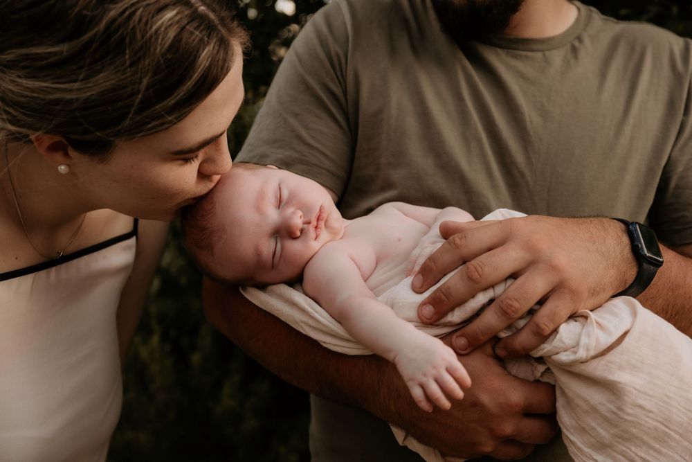 New mother tenderly kissing her sleeping newborn baby held in fathers arms outdoors.