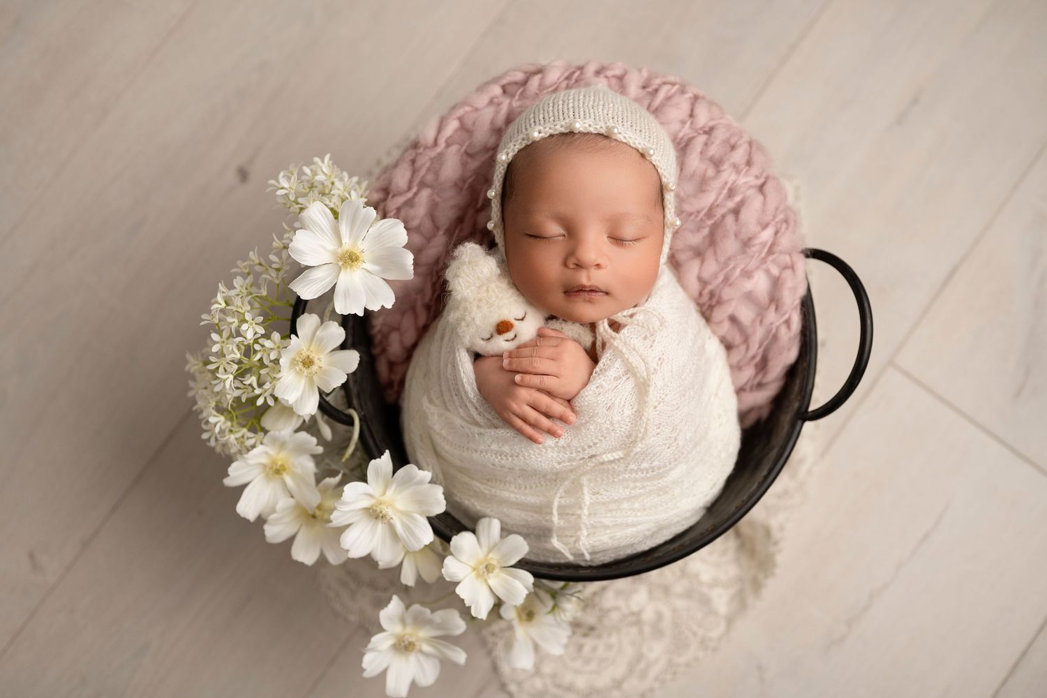 Newborn baby sleeping peacefully in a dark bowl with white flowers and pink knit wrap and bonnet.