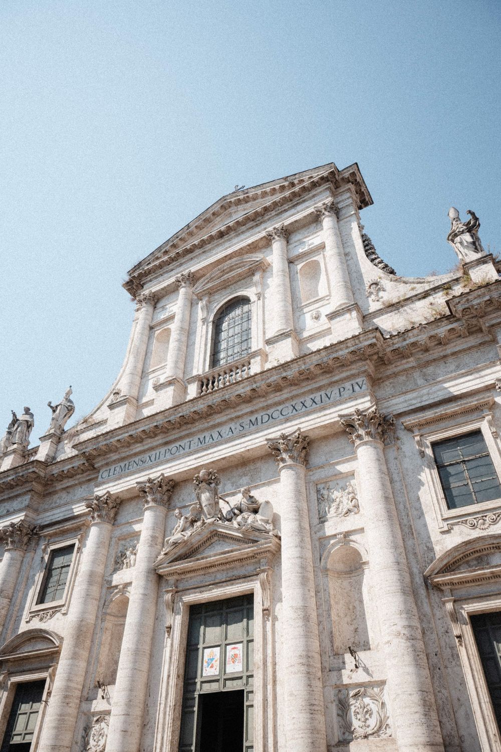 Baroque church facade with ornate columns, sculptures, and Latin inscription under a clear blue sky in Rome, Italy.