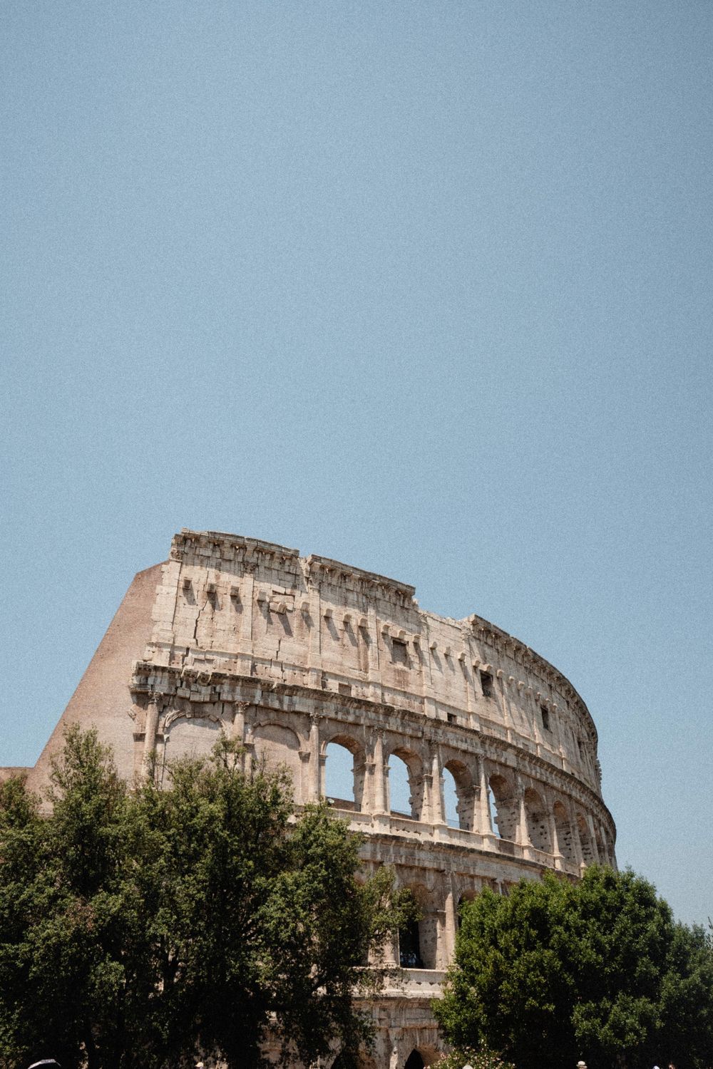 The ancient Roman Colosseum rises above green trees against a clear blue sky in Rome, Italy.