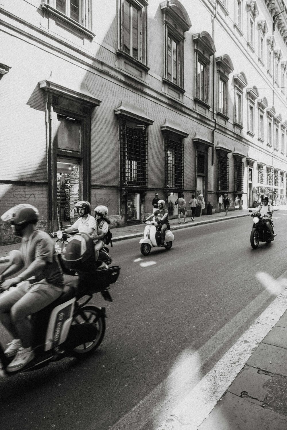 Black and white photo of motorcyclists and scooters riding down a classic European city street lined with historic buildings.